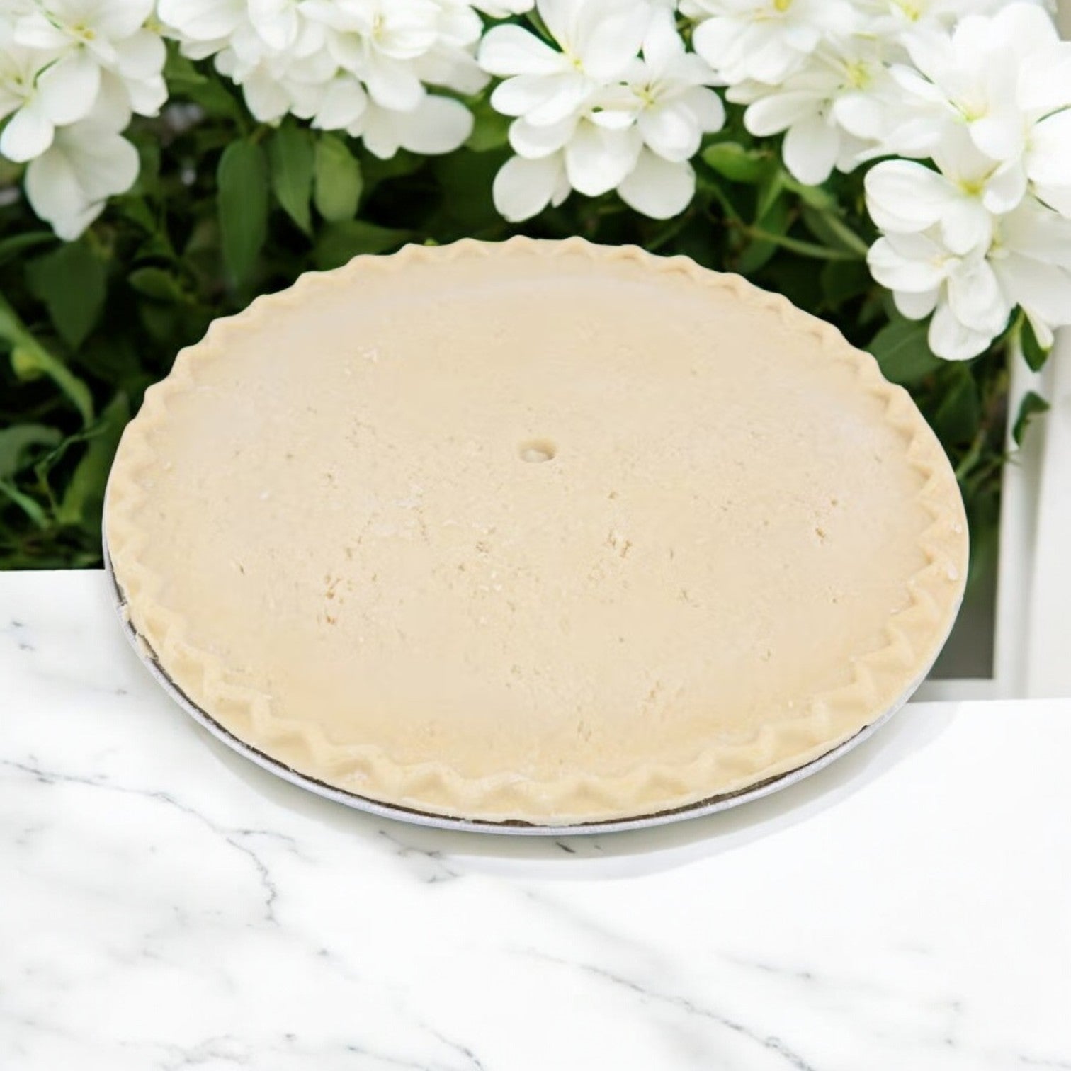 A 46 oz. round Chef Pierre Peach Pie with a crimped edge sits frozen and unbaked on a marble surface, set against white flowers, promising a sweet and juicy filling.