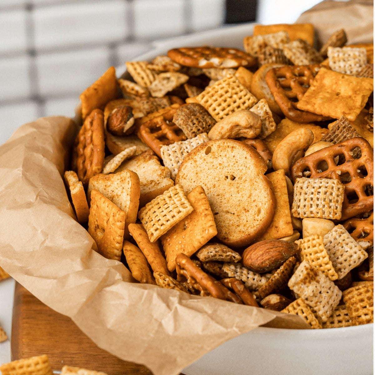 A bowl brimming with General Mills Chex Mix Traditional Savory Snack Mix, featuring pretzels, Chex cereal, bagel chips, and assorted seasoned crackers, rests on a piece of brown parchment paper. This snack is part of a Variety Pack including 42 individual bags.