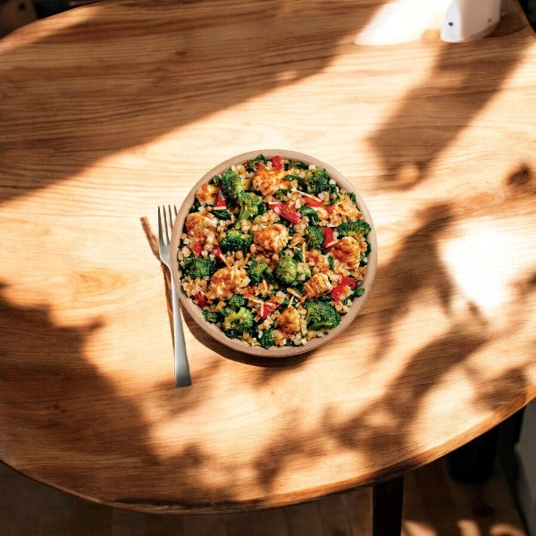 A bowl of salad with a fork on a wooden table. The salad contains kale, chopped vegetables, grilled chicken, and grains. Sunlight casts shadows across the table surface. This nutritious meal is reminiscent of the heartiness of Chicken Adobo but maintains the light and fresh appeal found in Healthy Choice Power Bowls Chicken Marinara With Riced Cauliflower by Healthy Choice.