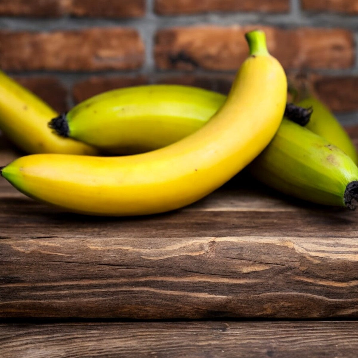 Four ripe Organic Bananas from Easy Lunches, three yellow and one green, are placed on a rustic wooden surface with a brick wall in the background. This 4lb bundle of natural goodness makes for nutritious snacks.