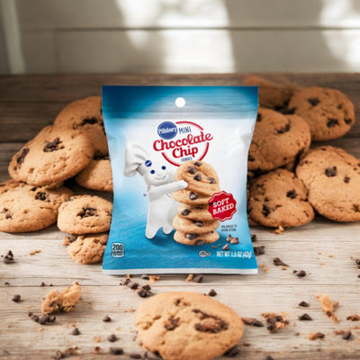 A package of Pillsbury Soft Baked Mini Chocolate Chips is placed among scattered soft-baked chocolate chip cookies on a wooden surface. The package features the Pillsbury mascot holding a stack of cookies.