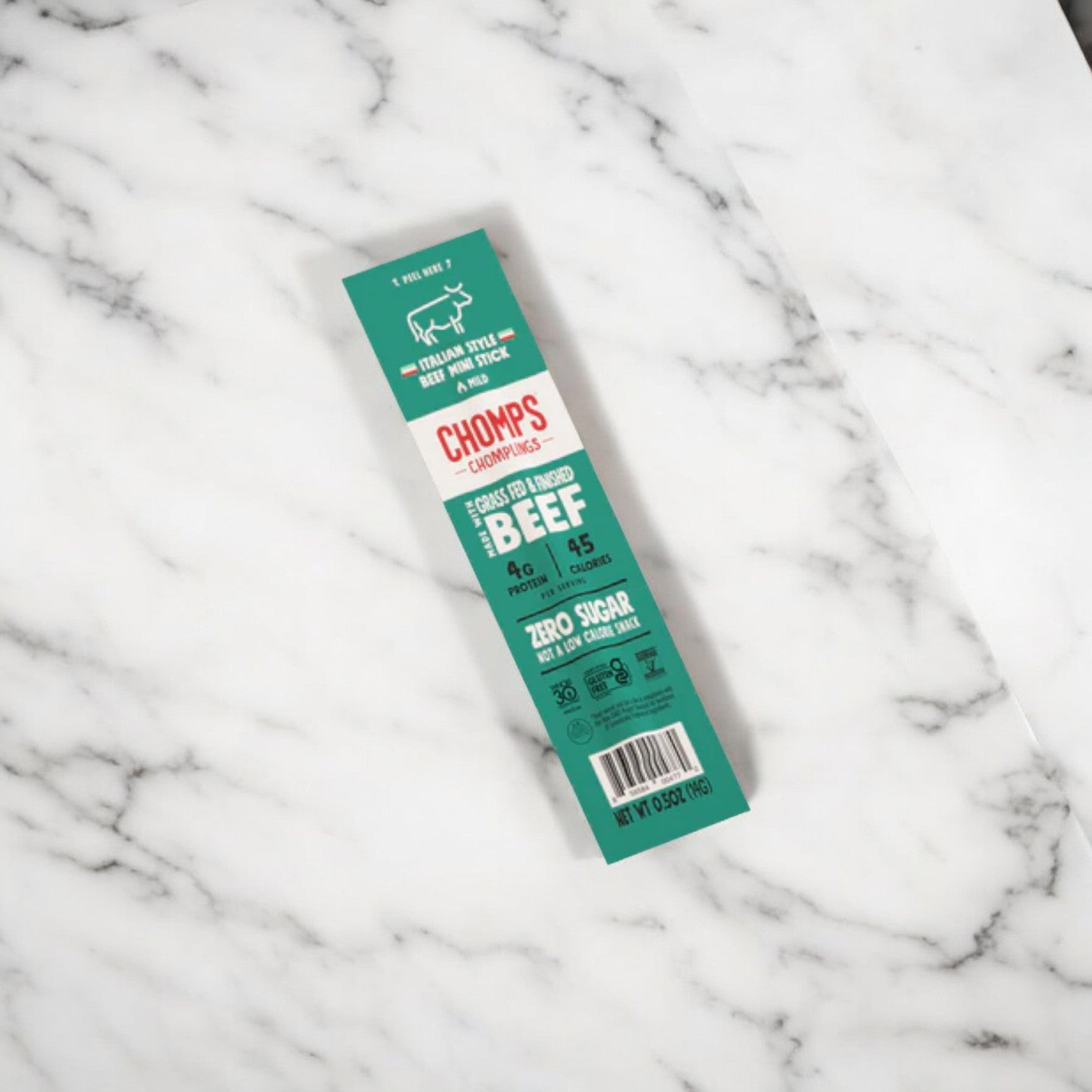 A green package of Chomps Mini Grass Fed Beef Jerky Meat Snack lies on a white marble surface. The gluten-free packaging highlights zero sugar, 9g of protein per serving, and its suitability for Keto diets.