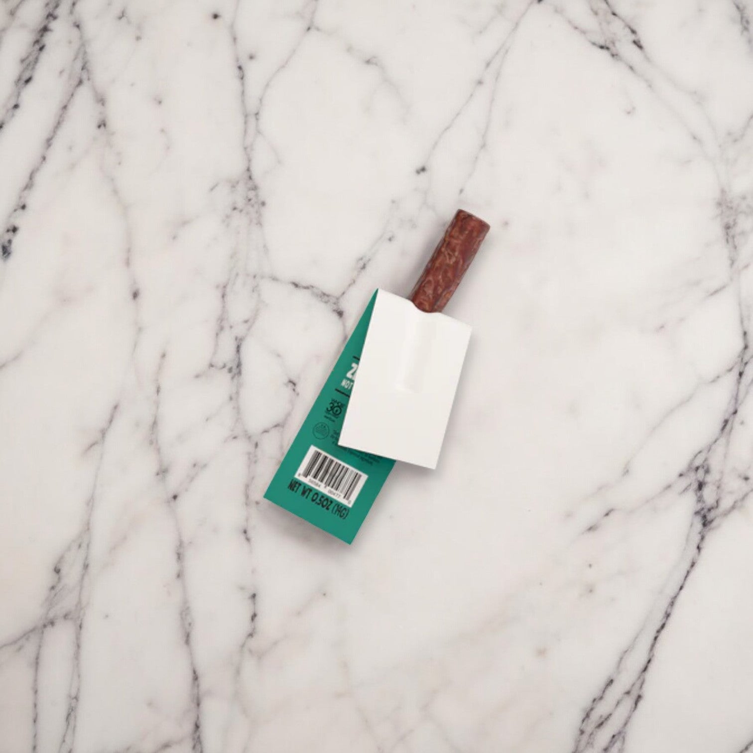 A small green and white package labeled "Chomps Mini Grass Fed Beef Jerky Meat Snack - 0.5 oz - 1 Count" with a barcode is partially covered by a white rectangular object with a brown handle, placed on a white marble surface.