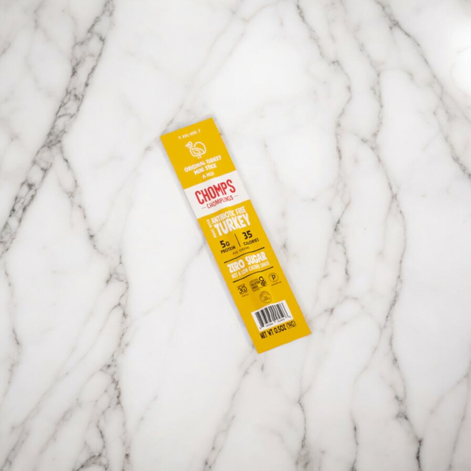 A Chomps Mini Antibiotic Free Original Turkey Chompling, in yellow packaging, rests on a white marble surface. The package states it contains 5g of protein per serving, zero sugar, and is Whole30 approved, paleo-friendly, and gluten-free. Savor this healthy meat snack made from sustainably sourced proteins.