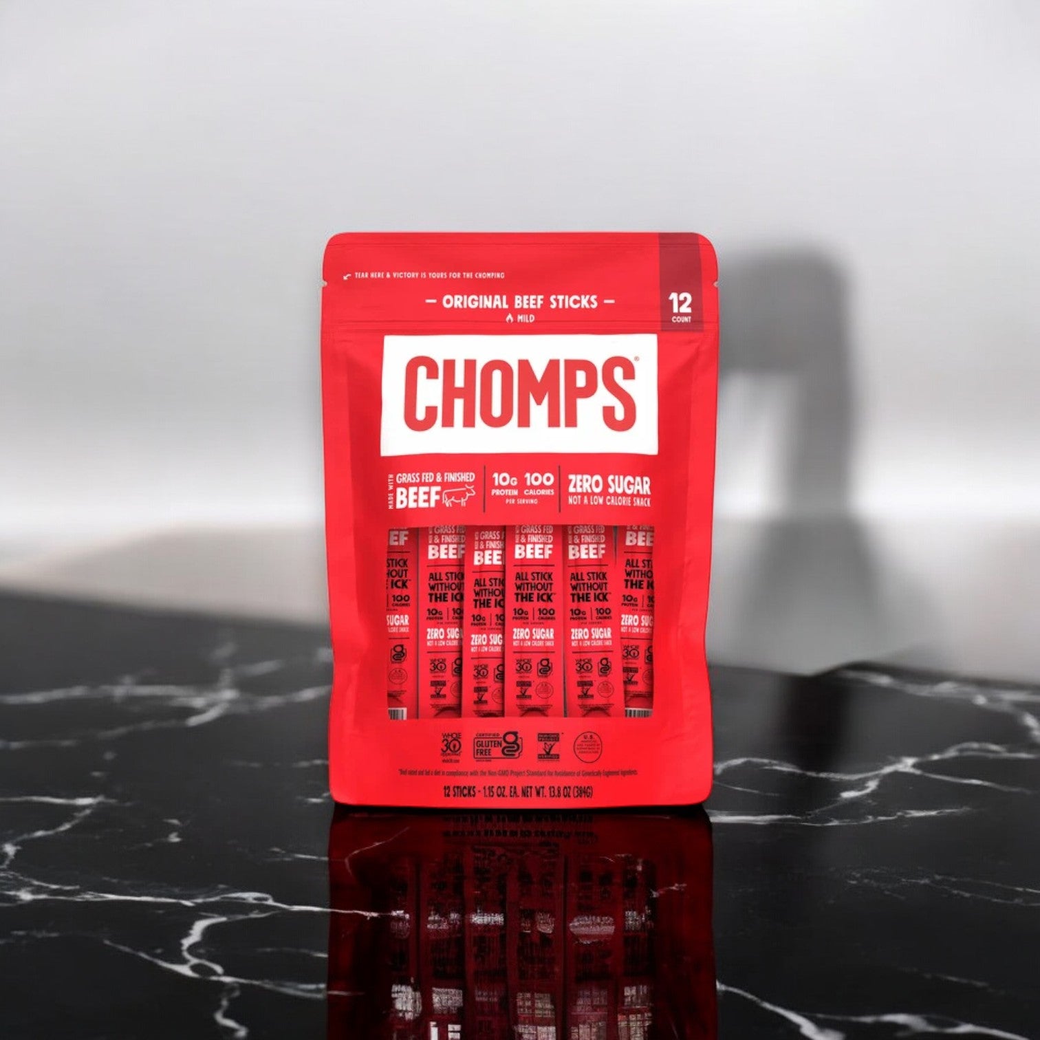 A package of Chomps Original Beef Sticks, Mild, containing 12 individually wrapped sticks, rests on a black marble surface. Each stick boasts 100 calories, 9g of protein, and zero sugar. Made with 100% grass-fed beef, this protein-packed snack is one you can feel great about.