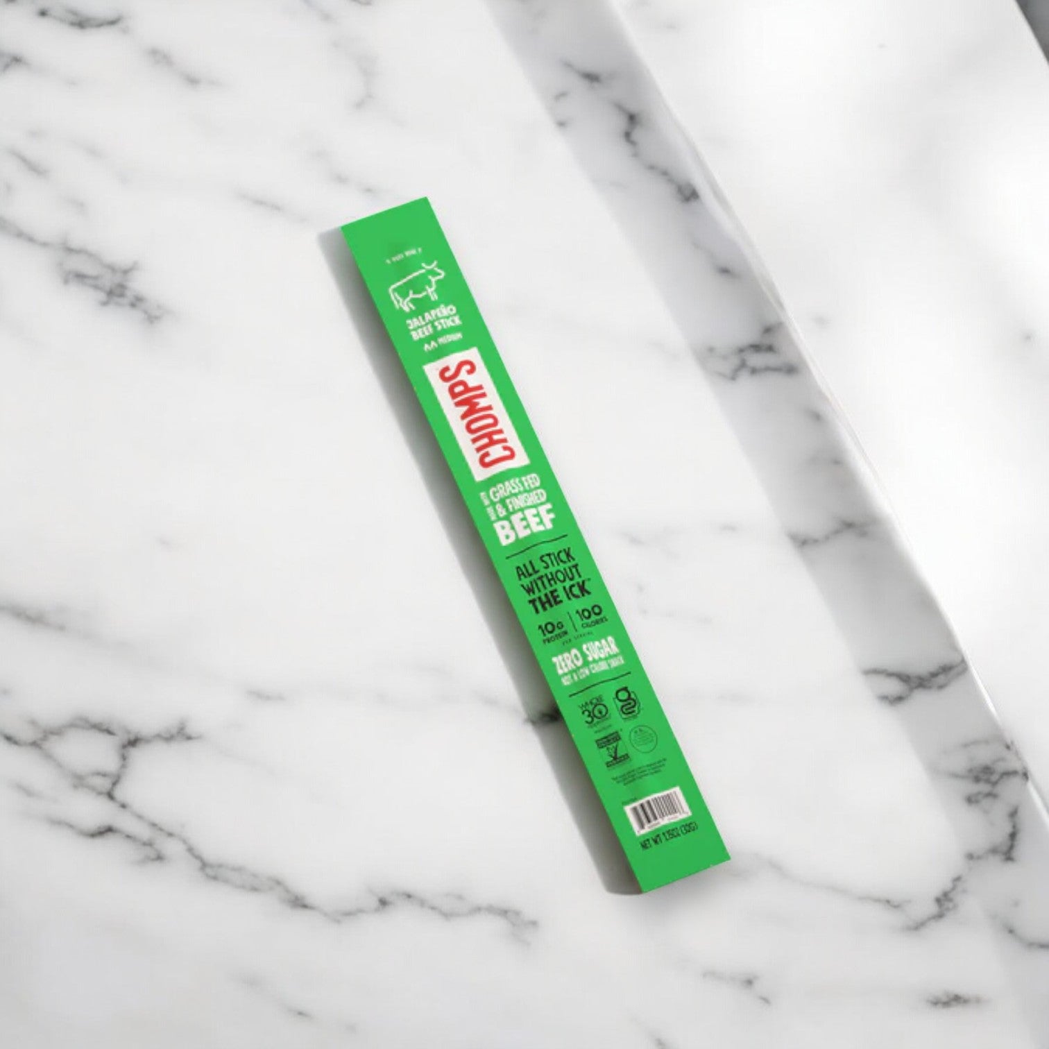 A green stick of Chomps Zero Sugar Jalapeno Beef Jerky Sticks -1.15 oz -1 Count lies on a white marbled surface. The packaging highlights its attributes such as being grass-fed, non-GMO, keto-friendly, gluten-free, and only 100 calories.