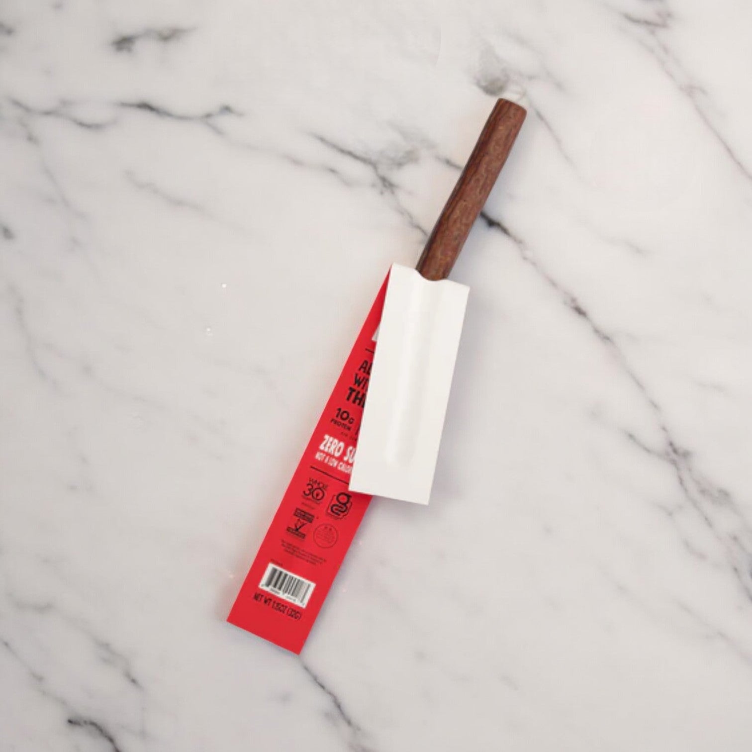 A metal utensil with a wooden handle is partially wrapped in red and white packaging, placed on a white marble surface—perfect for slicing into some delicious Chomps Zero Sugar Original Beef Jerky Sticks from Chomps.