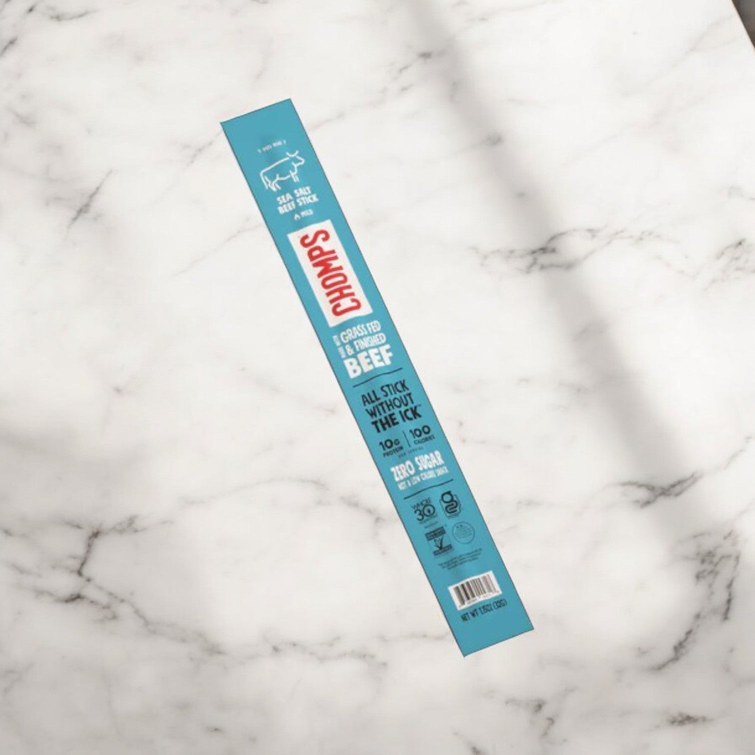 A Chomps Grass-Fed and Finished Sea Salt Beef Jerky Meat Snack in its blue packaging lies on a white marble surface. The packaging highlights key features such as zero sugar, 1g carb, 9g protein, and non-GMO ingredients, all while boasting sustainably sourced proteins.