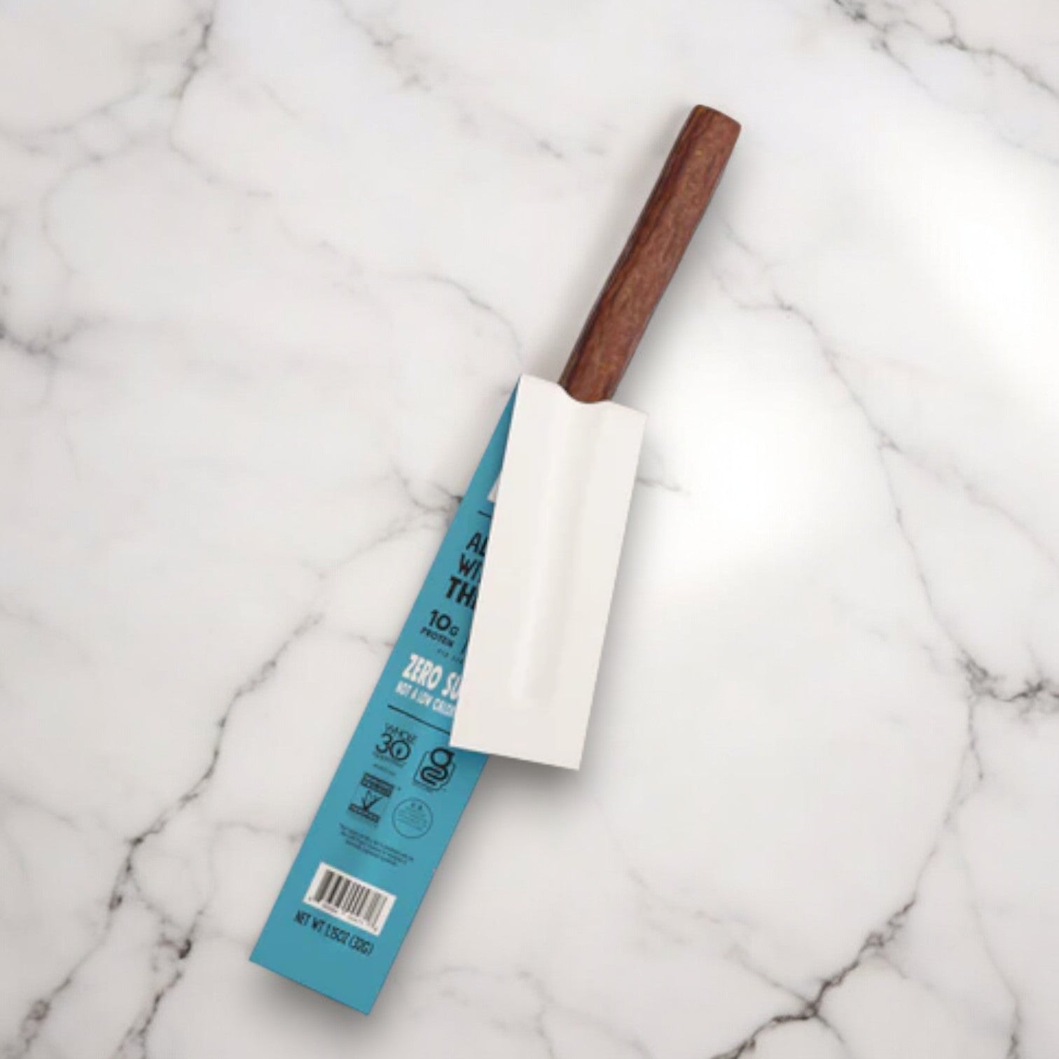 A Chomps Grass-Fed and Finished Sea Salt Beef Jerky Meat Snack, 1.15 oz Stick -1 Count, featuring a wooden handle and enclosed in blue packaging, is elegantly displayed on a white marble surface. The packaging showcases text and various icons that proudly highlight its sustainably sourced proteins.