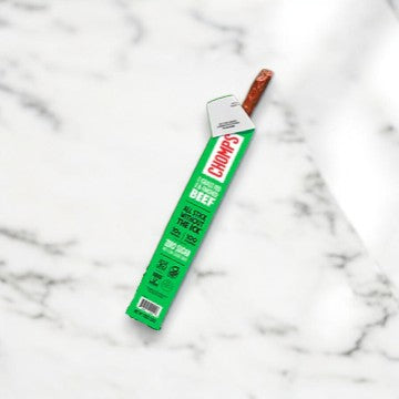 A Chomps Zero Sugar Jalapeno Beef Jerky Stick, weighing 1.15 oz, is placed on a marble surface. The packaging, featuring green and white with red text, proudly highlights its grass-fed beef origins, making it an ideal keto-friendly snack.