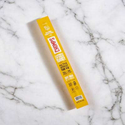 A yellow package of Chomps Original Turkey Jerky Meat Snack Stick, 1.15 oz Stick - 1 Count is placed on a marble surface. The packaging highlights that it contains 12g of protein, 30 calories, zero sugar, and is gluten-free. These healthy meat snacks from Chomps are also Whole30 Approved for your dietary needs.