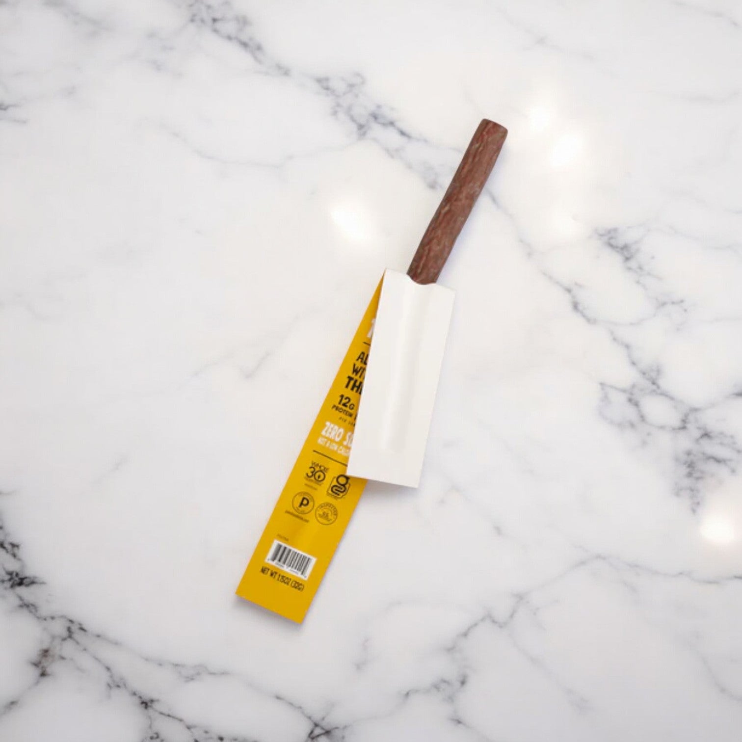 A yellow plastic package with a barcode and text is partially open on a white marble surface, revealing the brown handle of what appears to be a Chomps Original Turkey Jerky Meat Snack Stick inside.