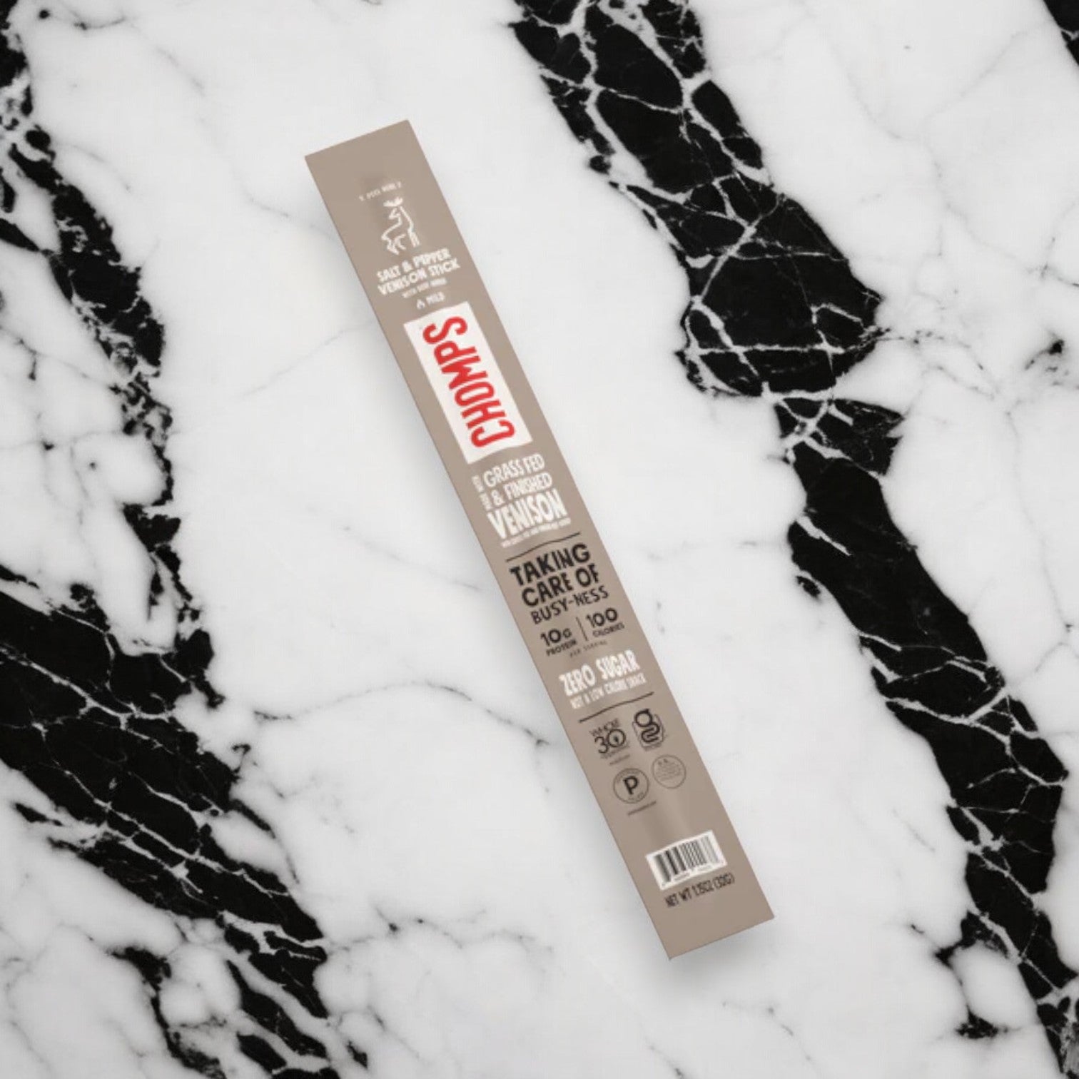 A Chomps Venison Stick - Pepper - Salt - 1.15 oz is placed on a black and white marble surface. The packaging highlights "Taking care of our kids and the planet" and boasts "Zero Sugar," "100 Calories," as well as being a protein-packed snack option.