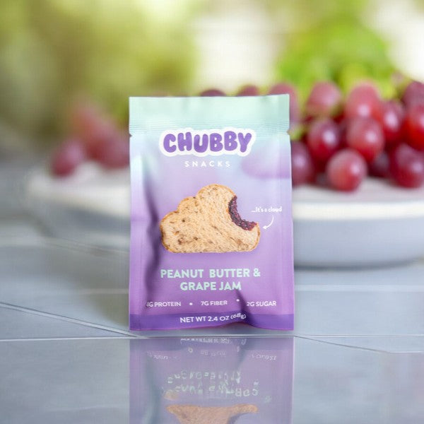 A package of Chubby Snacks - Peanut Butter & Grape Jam (2 oz, 1 Count) is in focus with a plate of grapes in the soft-focus background, showcasing a protein snack made from wholesome ingredients.