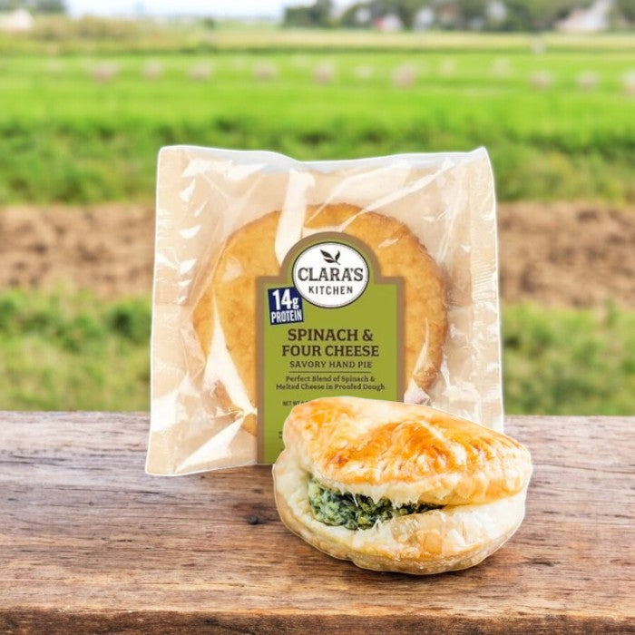 A Clara’s Kitchen Four Cheese Spinach Handpie – 6 oz., set on wood with fields beyond, entices with its vegetarian-friendly gourmet charm, offering a delightful experience from package to plate.