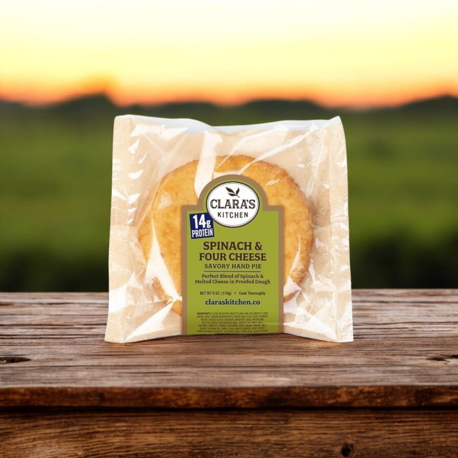 A Clara’s Kitchen Four Cheese Spinach Handpie – 6 oz. with a flaky crust rests on a wooden surface against a blurred green landscape.