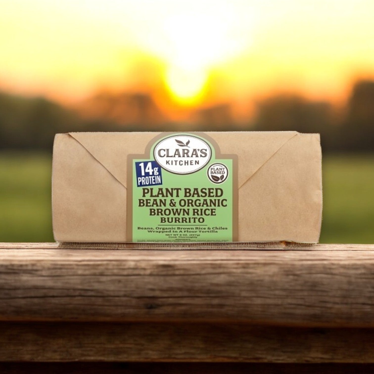 A Clara's Kitchen Individually Packaged Vegan Bean & Cheese Burrito, 8 oz., rests on a wooden surface, capturing the essence of a breakfast sandwich enjoyed at sunrise, with a blurred sunset behind it.