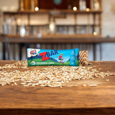 A Clif Kid Zbar Organic Iced Oatmeal Cookie Snack Bar, 1.27 oz, from Clif Bar, is placed on a wooden table with scattered oats and a blurred background of shelves in a kitchen.