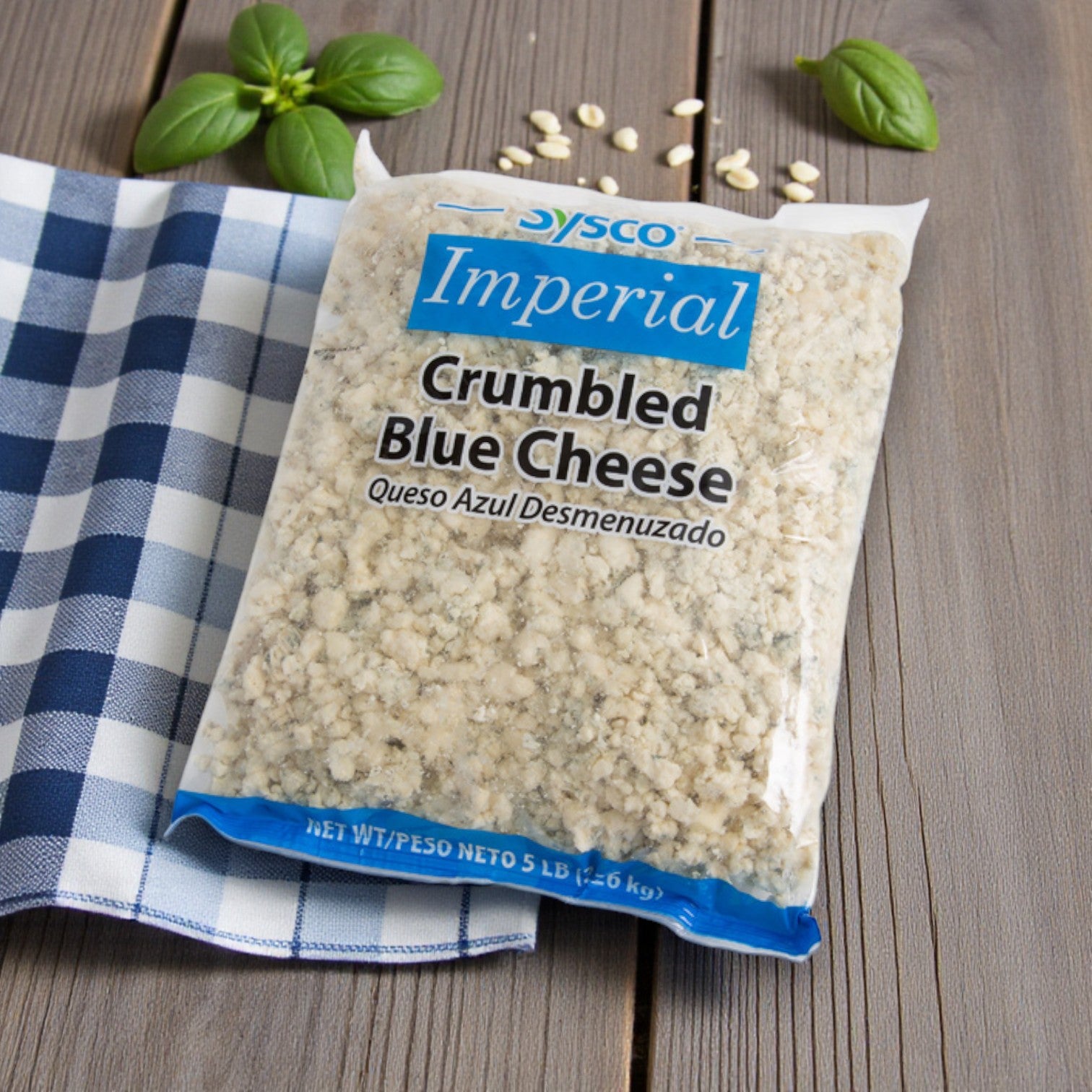 A package of Sysco Imperial – Blue Cheese Crumbles (2/5 lb) rests on a wooden surface with a blue checkered cloth, basil, and white beans—ideal for foodservice kitchens seeking gourmet ingredients.