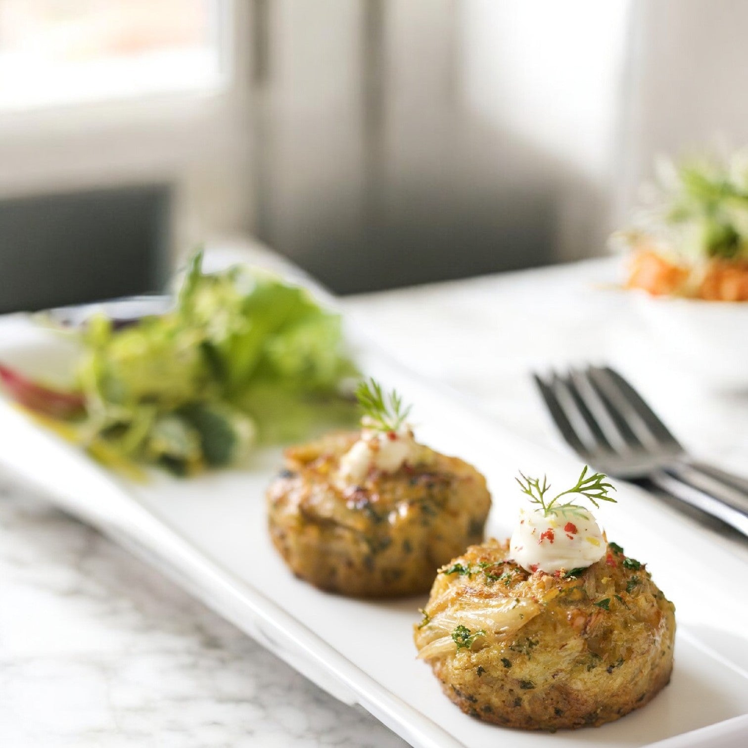 Two Crab Cake Chesapeakes, crafted with Phillips Foods' wild-caught Jumbo Pasteurized Asia Crab Meat, are topped with a dollop of sauce and fresh dill on a rectangular plate, served alongside a crisp side salad.