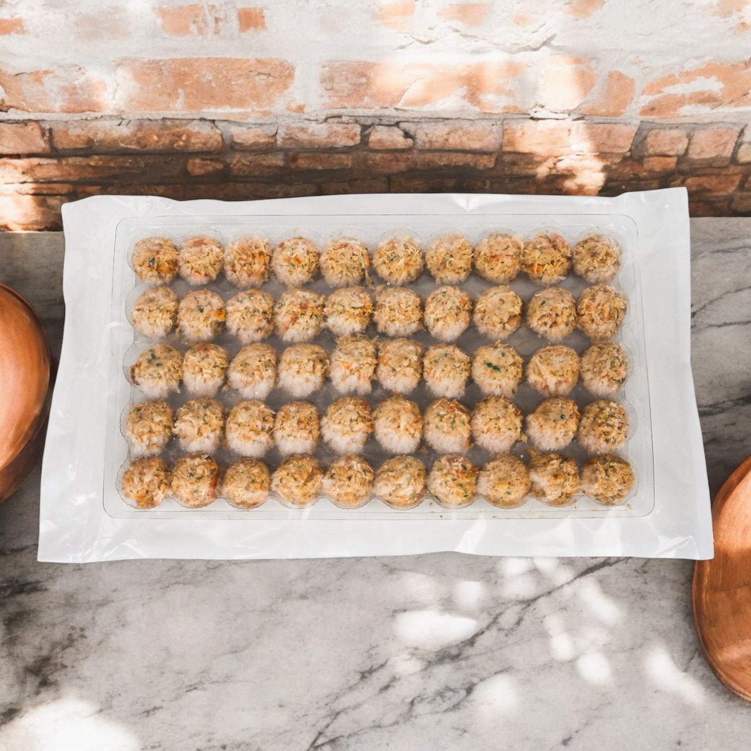 A sealed tray of 45 evenly spaced mini crab cakes from Phillips Foods, each made with sustainable sourcing, is set on a marble surface against a brick wall background.