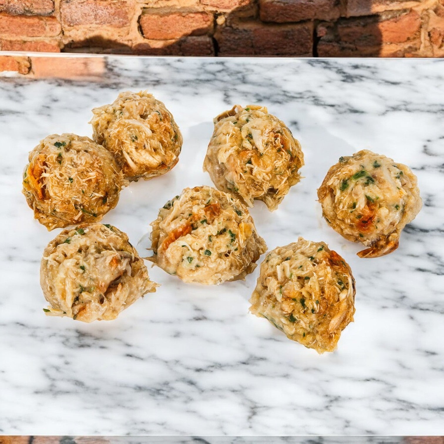 Seven mini crab cakes with herbs and jumbo lump crab meat rest on a marble surface, set against a brick wall background. These are from the Phillip Foods Crab Cake Mini Claw collection, .75 oz., 1 Case containing 100 pieces by Phillips Foods.