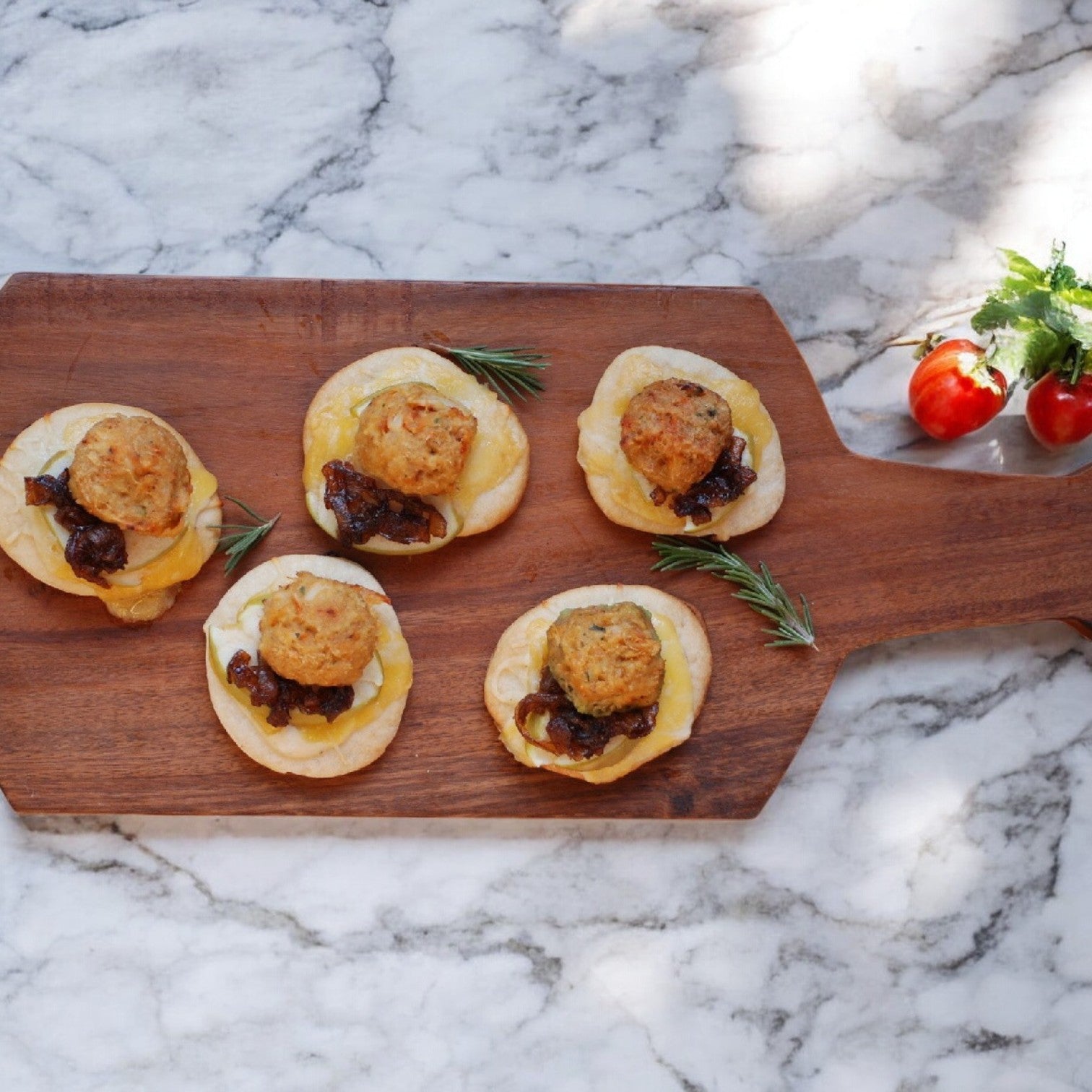 Five Phillips Foods Crab Cake Mini Claws, featuring cheese, caramelized onions, crab meat, and a small patty on round bread, are elegantly arranged on a wooden board. A rosemary sprig and cherry tomatoes rest on the nearby marble surface.