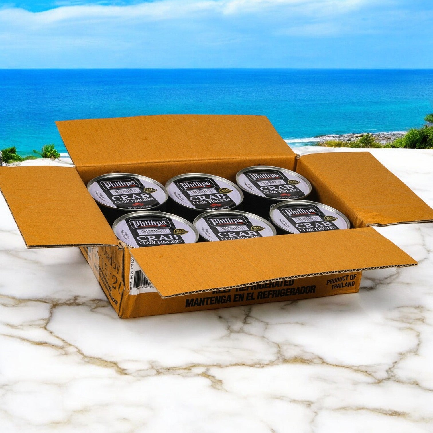 An open cardboard box displays several cans of Phillips Foods Crab Claw Finger Pasteurized Asia on a marble surface, set against a scenic ocean backdrop. These products promise authentic flavors and offer versatility for crafting appetizers ideal for any occasion.