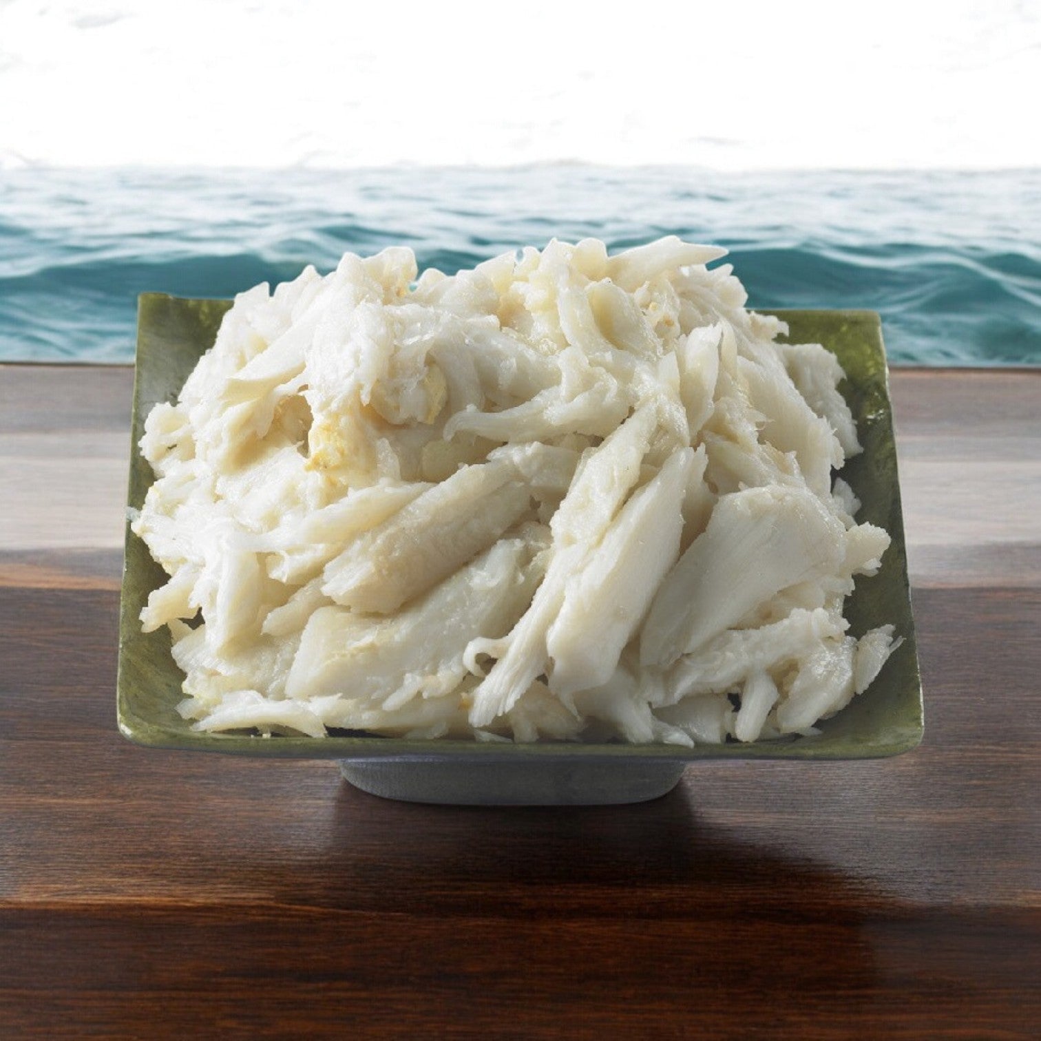 A square green bowl filled with white, shredded sustainable crab meat from Phillips Foods sits gracefully on a wooden table, against the backdrop of a serene water body.