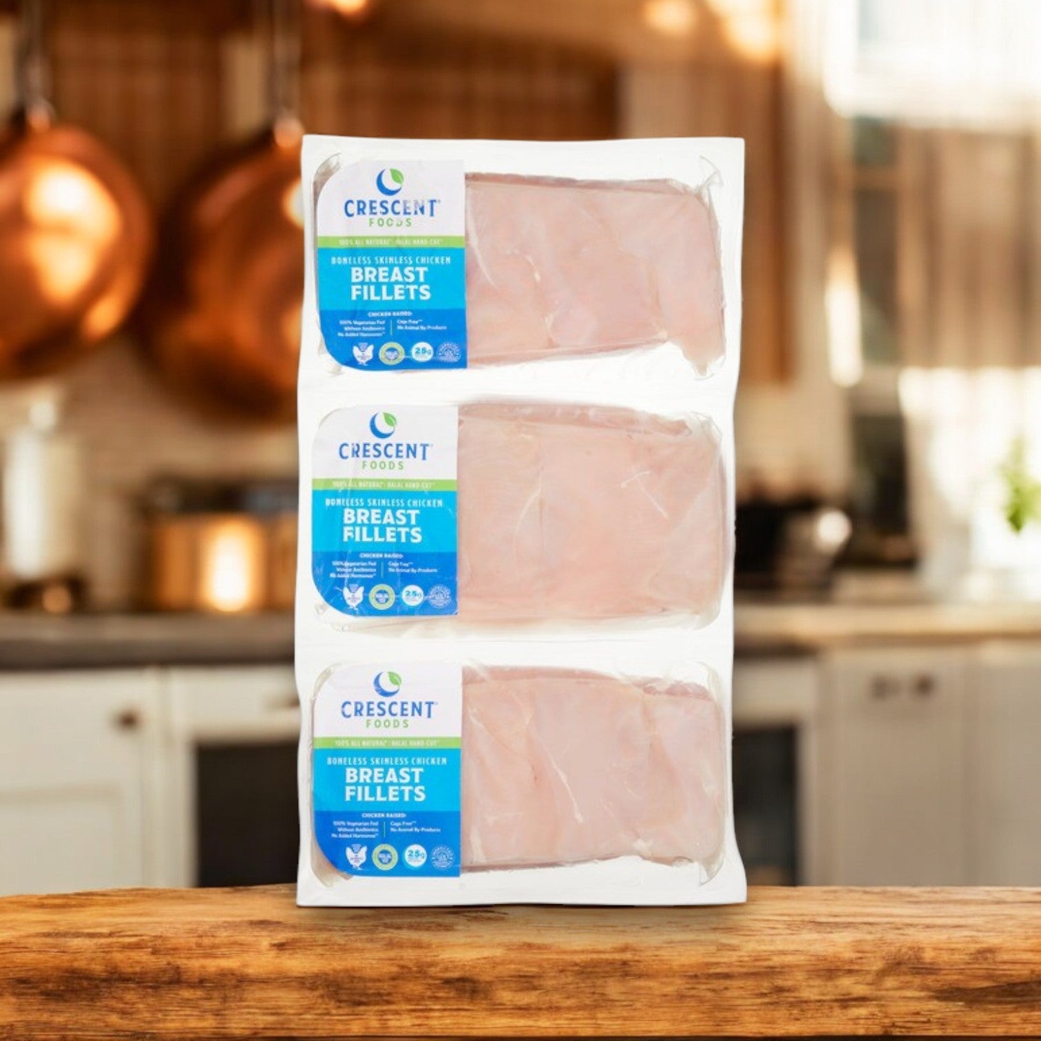 Three packs of Crescent Foods Halal Chicken Breast, Hand-Cut, Boneless & Skinless (Approx. 4 lbs) are stacked on a kitchen counter with copper pots gleaming in the background.