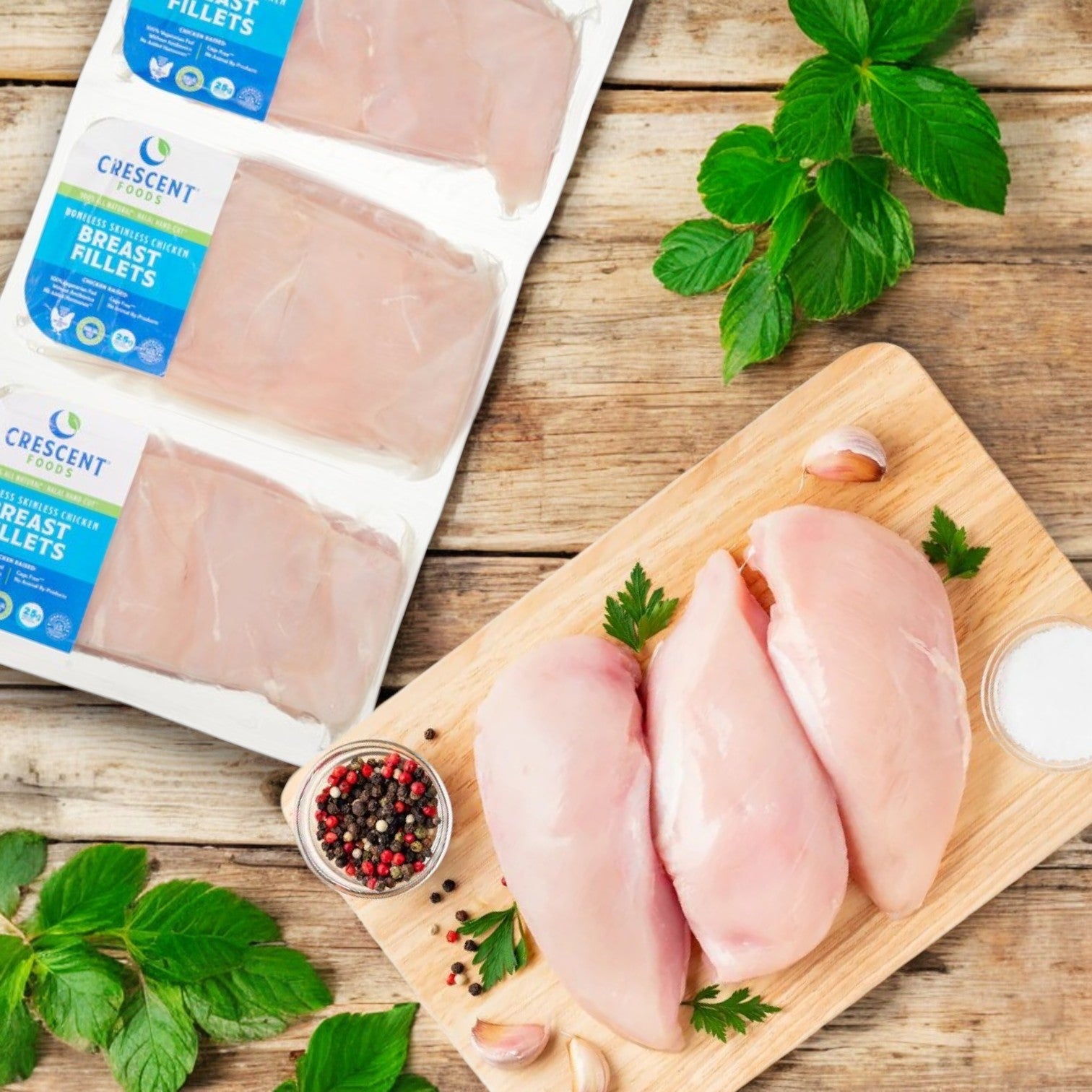 Three raw boneless, skinless chicken breasts sit on a wooden board with garlic, salt, parsley, and peppercorns. Nearby are packed fillets labeled "Crescent Foods Halal Chicken Breast," capturing the premium quality of Crescent Foods. Mint leaves scattered around complete the scene.