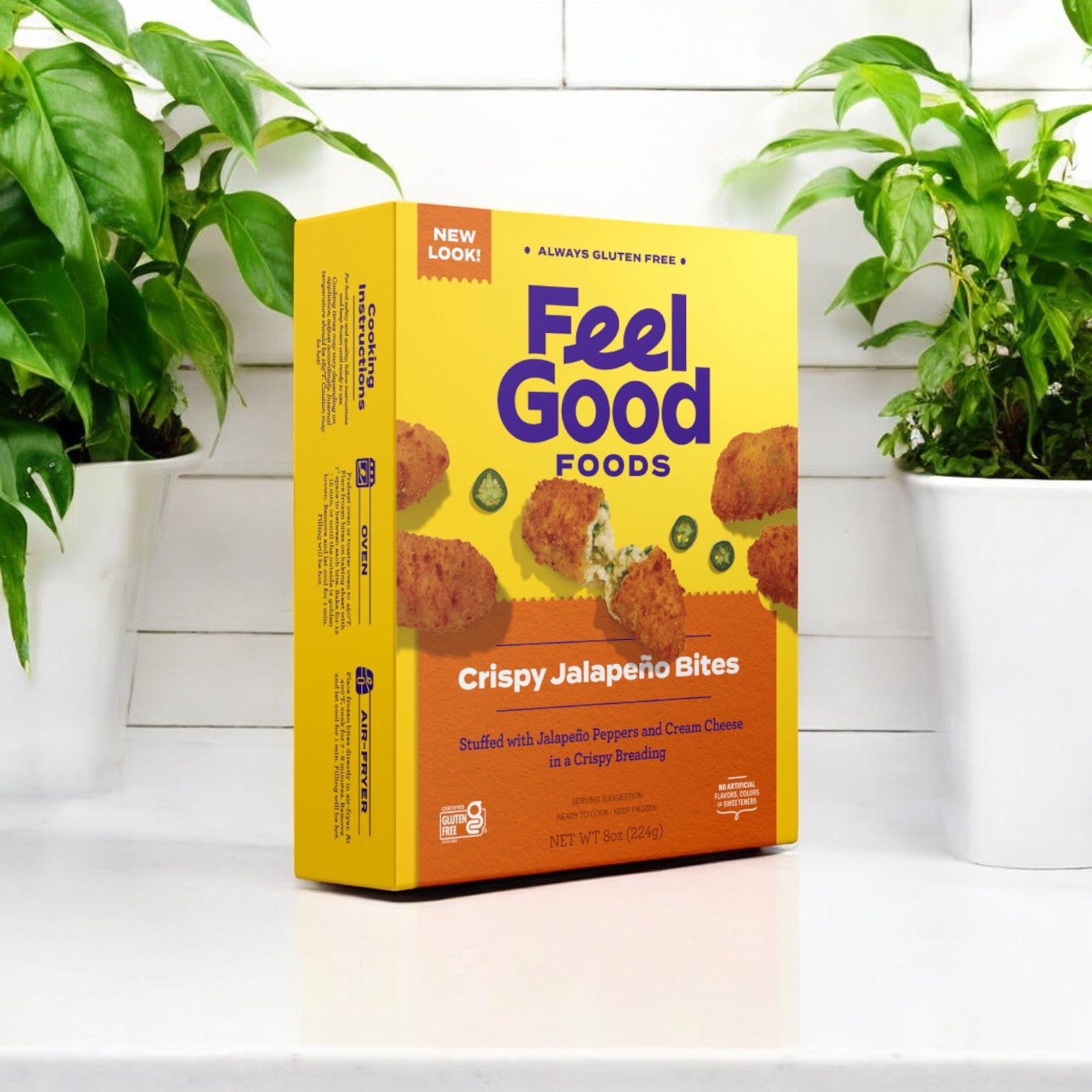 An 8 oz box of Feel Good Foods Crispy Jalapeno Bites rests on a white countertop, surrounded by two potted plants. These bites, with their spicy jalapeño filling and irresistible crunch, offer a delightful snacking experience.