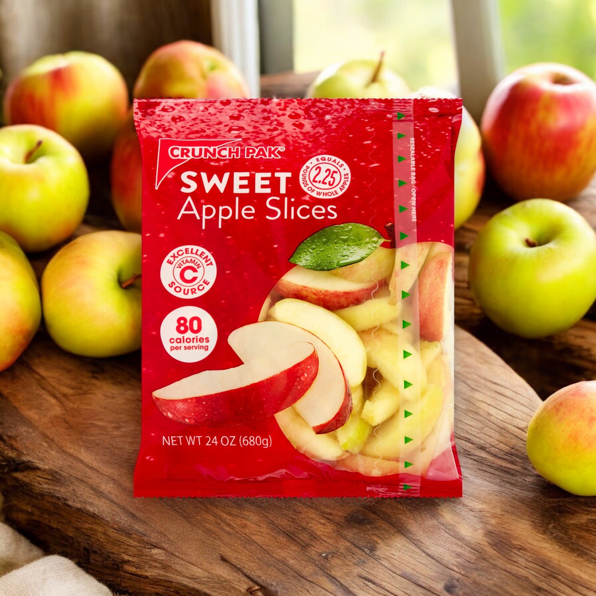 A package of Crunch Pak Fresh Sweet Apple Slices, Family Size (24oz Resealable Bag) is displayed on a wooden table, surrounded by whole apples. The packaging, perfect for on-the-go snacks, indicates 80 calories per serving.