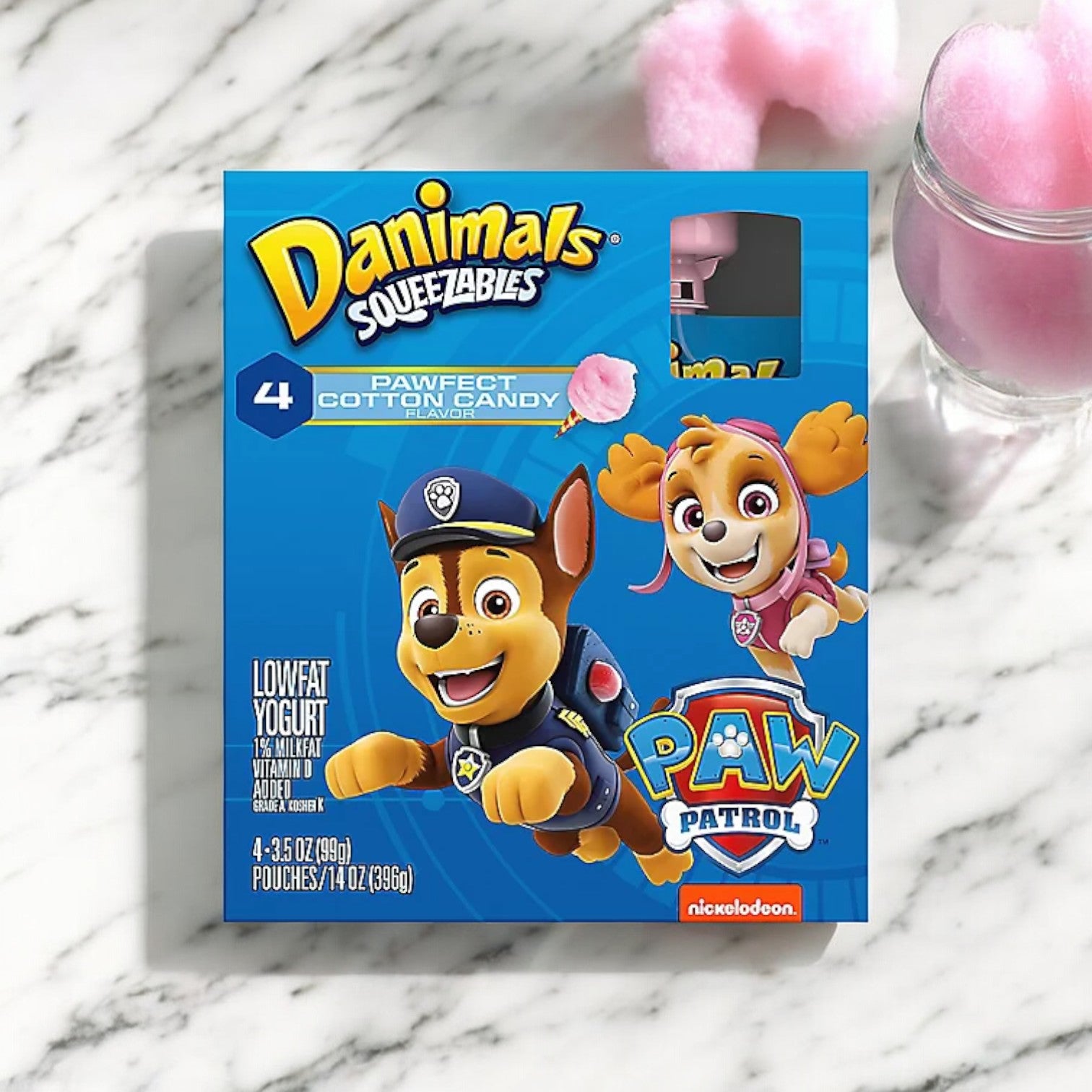 A blue box of Easy Lunches Danimals Cotton Candy Squeezable Yogurt featuring "PAW Patrol" characters. Contains 4 pouches of lowfat, nutrient-rich yogurt with a delightful cotton candy flavor. The box is placed on a marble surface next to a cotton candy serving.