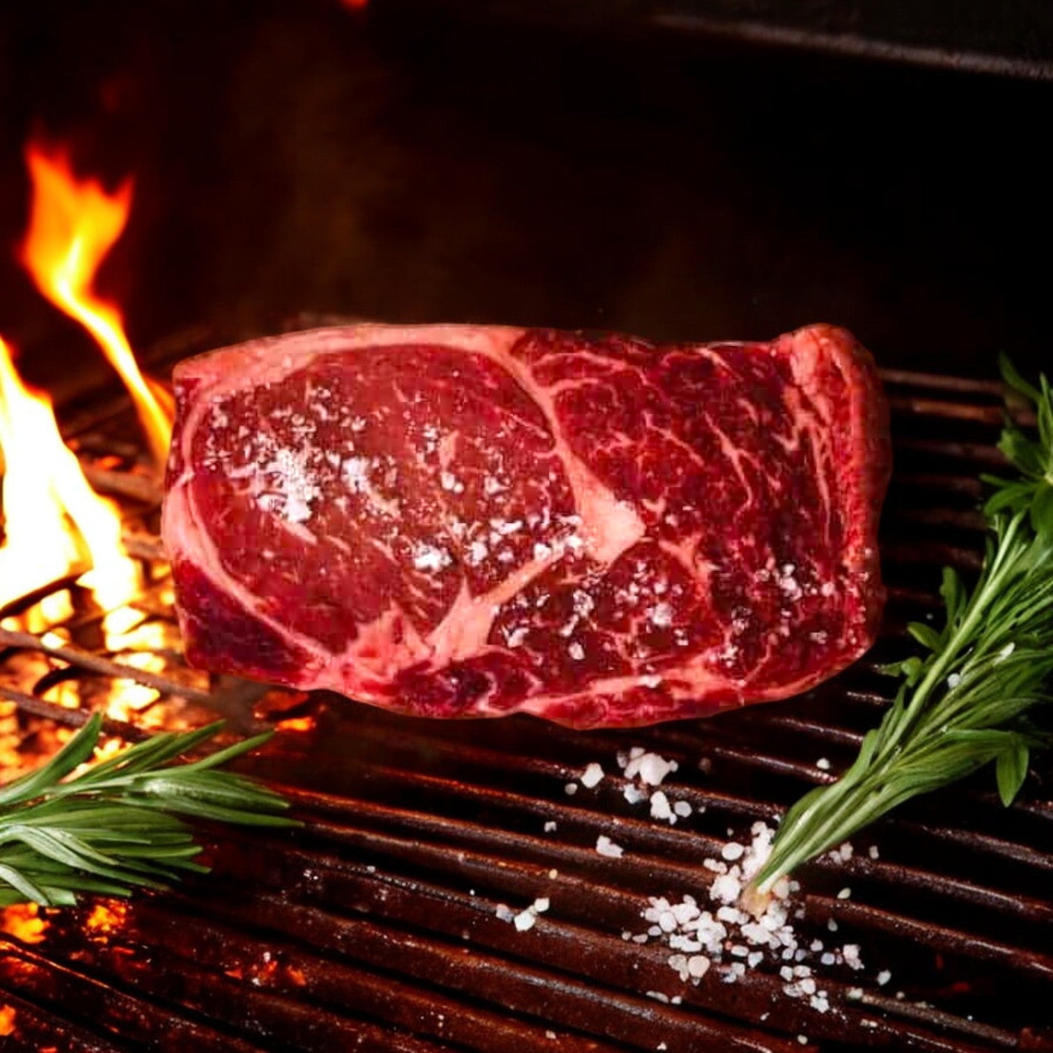 A 16 oz Steegs Cut Delmonico Ribeye from Mino Prime Steaks sizzles on the grill, seasoned with salt, surrounded by fresh rosemary sprigs, as an open flame flickers in the background.