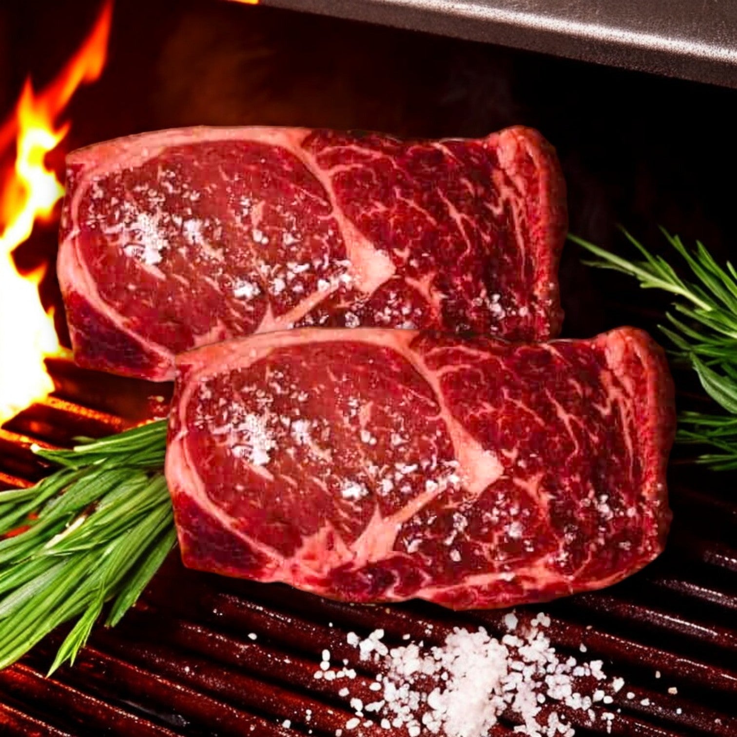 Two seasoned steaks from the Mino Prime Steaks - Top Seller Box sizzle on the grill, flames dancing behind them. Garnished with rosemary and coarse salt, it's a tempting sight for steak lovers craving premium meats.