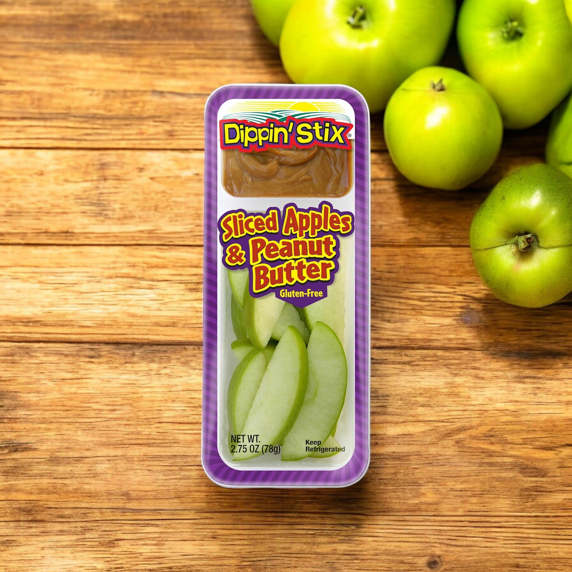 A container of Dippin' Stix Sliced Apples & Peanut Butter - 2.75 oz - 1 Count from Dippin' Stix rests on a wooden surface, with green apples in the background enhancing its appeal. This nutritious snack is gluten-free and weighs 2.75 oz (78g).