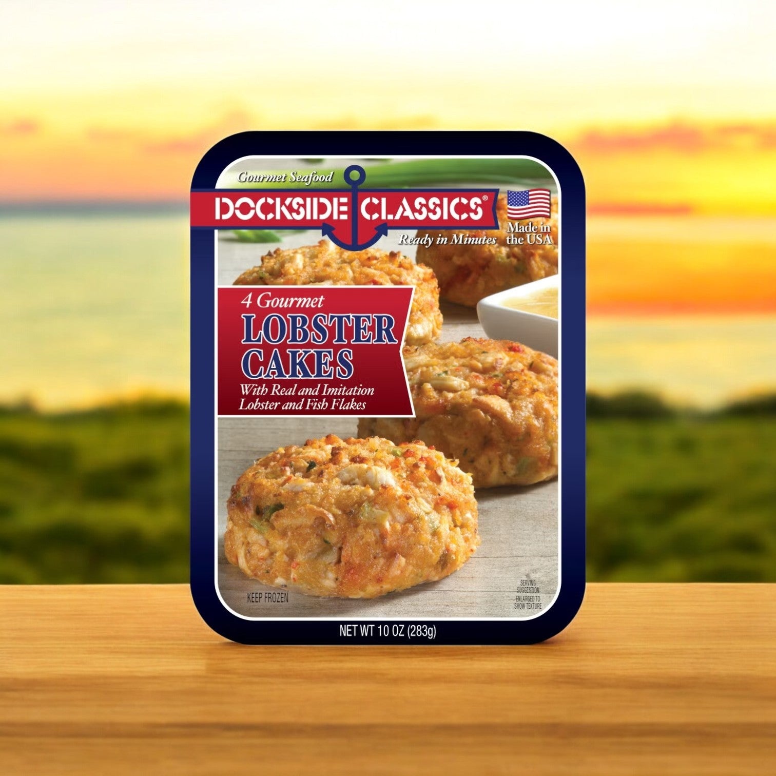 Front view of Dockside Classics Premium Lobster Cakes packaging against a sunset backdrop, highlighting "4 Gourmet Lobster Cakes." Made with real and imitation lobster along with fish flakes, these cakes offer a gourmet dining experience featuring high-quality ingredients.