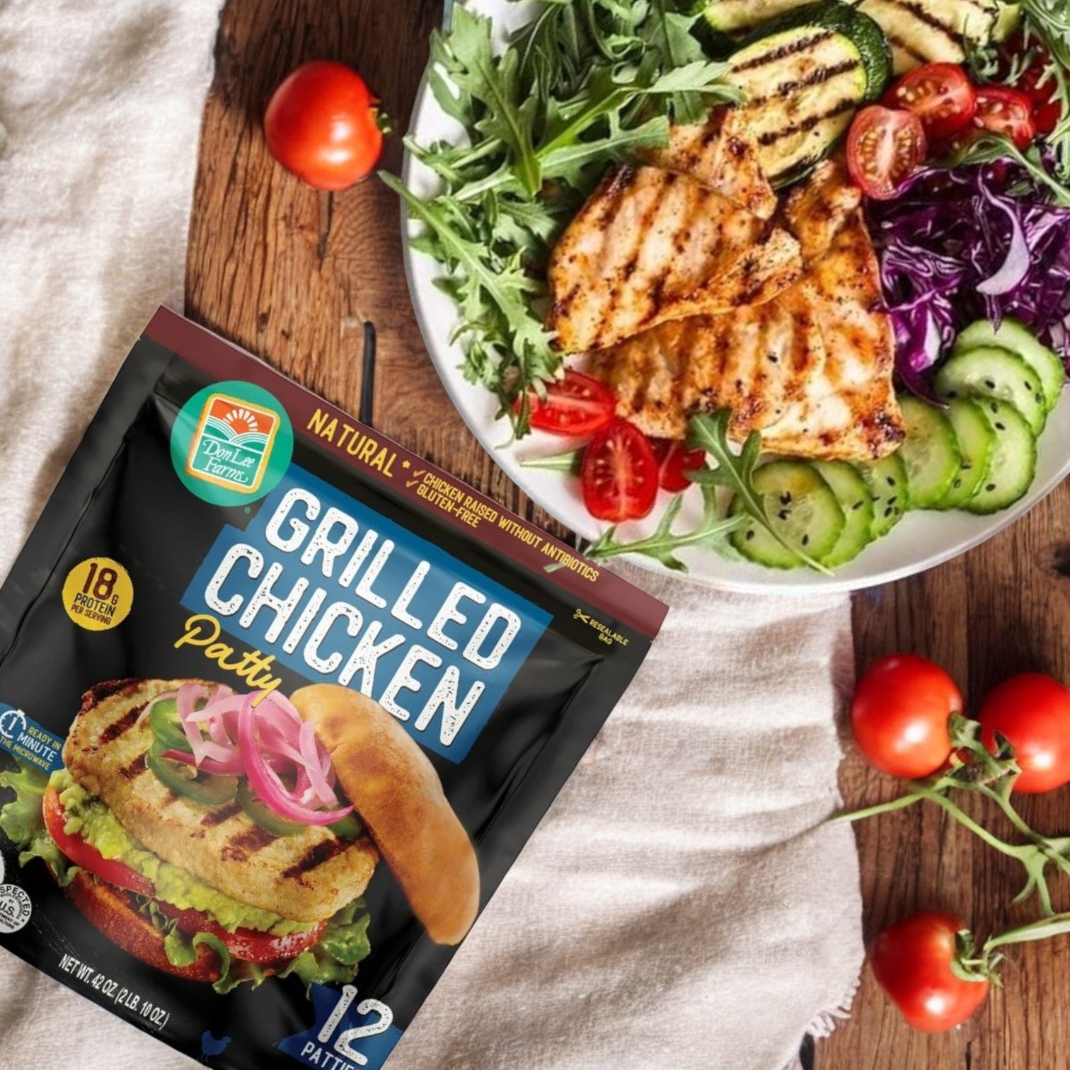 A plate of grilled chicken salad with cucumbers and greens sits next to a bag of Don Lee Farms Chicken Patties, 12-Count – Delicious, All-Natural & Fully Cooked, with cherry tomatoes scattered on a wooden table.