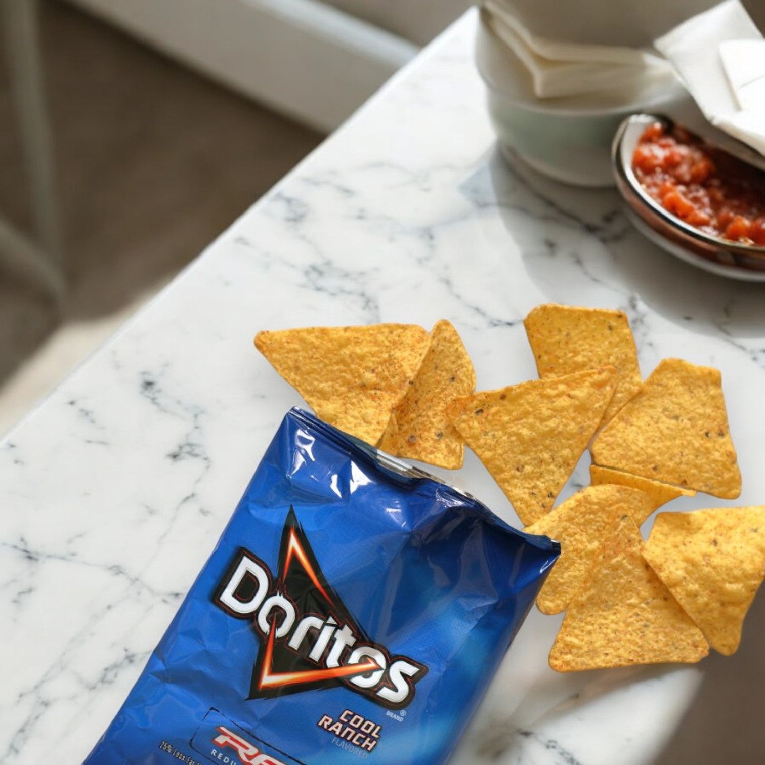 A marble table with a single-serving bag of Frito Lay Doritos Cool Ranch Flavored Tortilla Chips, 1 oz Bags - 1 Count, and some chips scattered beside it. A bowl of salsa and napkins are visible in the background, making it perfect for on-the-go snacking.