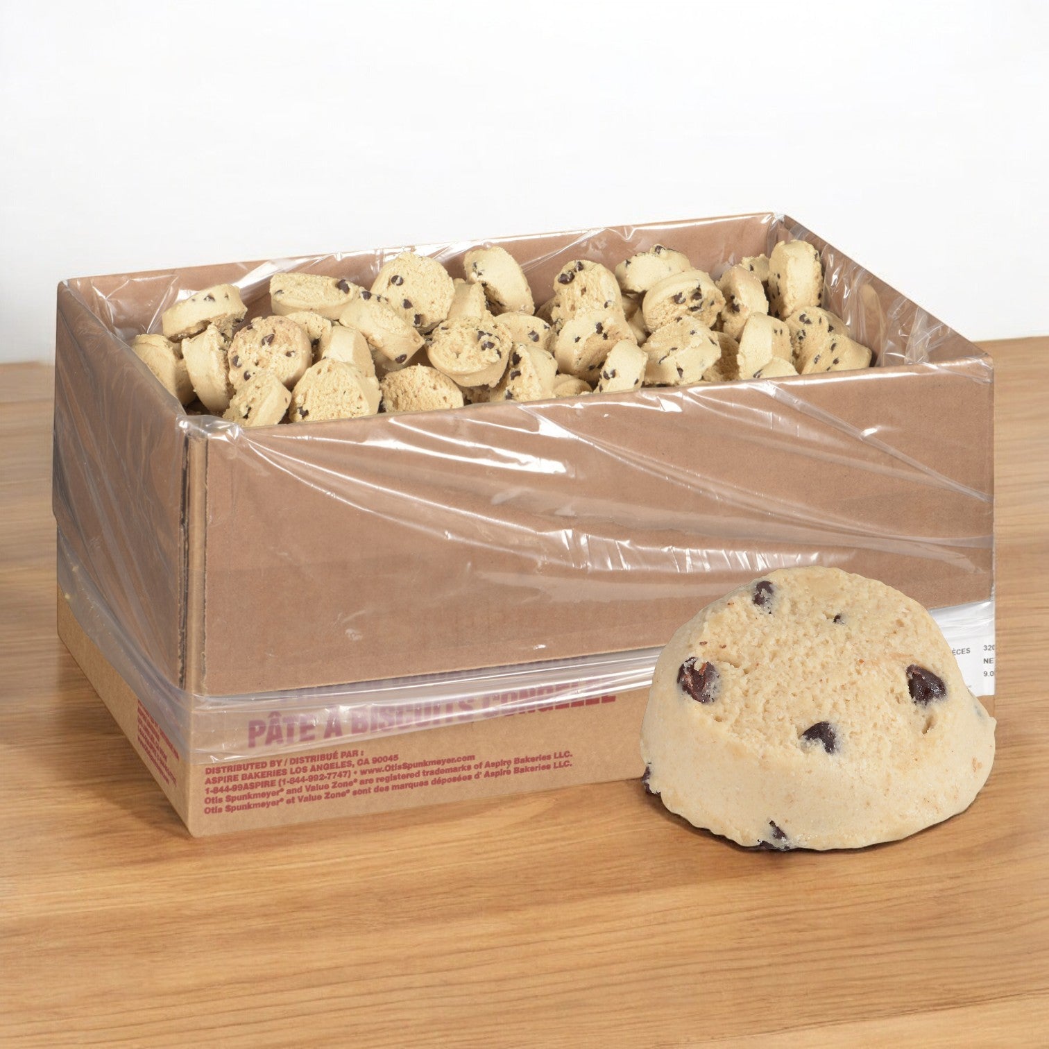 A wooden surface features a cardboard box of Otis Spunkmeyer Dough Cookie Chocolate Chip encased in a plastic liner. Beside the box is a large, rounded piece of edible chocolate chip cookie dough studded with visible chips from the Otis Spunkmeyer brand.