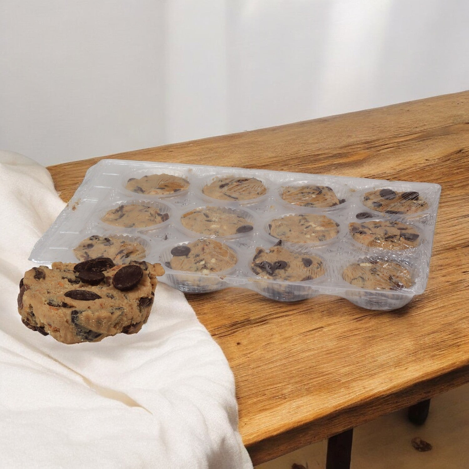 On a wooden table sits a tray of Sweet Street Dough Cookie Chocolate Chunk portions, with one piece placed on a white cloth nearby.