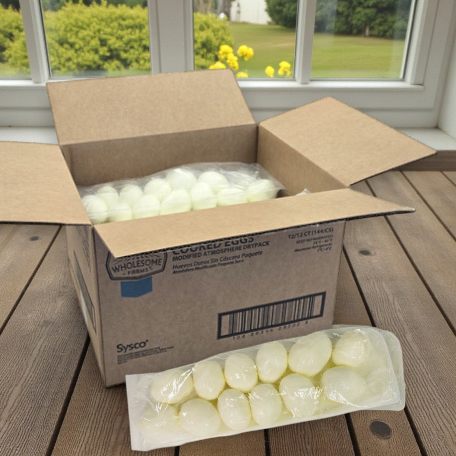 A box of Sysco Wholesome Farms Classic Eggs, Hardcooked & Peeled, Dry Pack – 12/12 ct, contains vacuum-sealed packs of eggs and rests on a wooden surface by a window.
