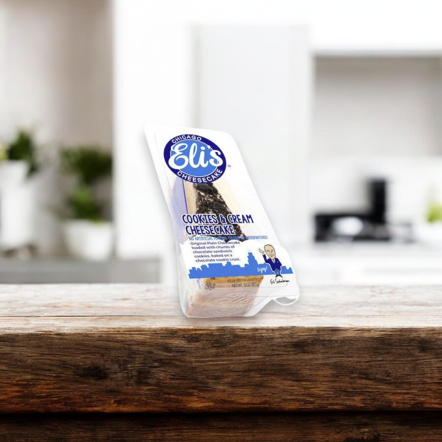 A single serve slice of Eli's Cookies and Cream Cheesecake, 3 oz., rests on a wooden counter with a blurry kitchen background.