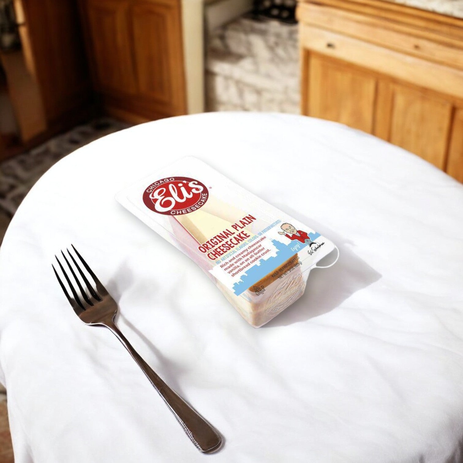 A sealed package of Eli's Cheesecake Original Plain Single Serve, 3 oz. - 1 Count is placed on a white surface next to a fork, with wooden kitchen cabinets in the background.