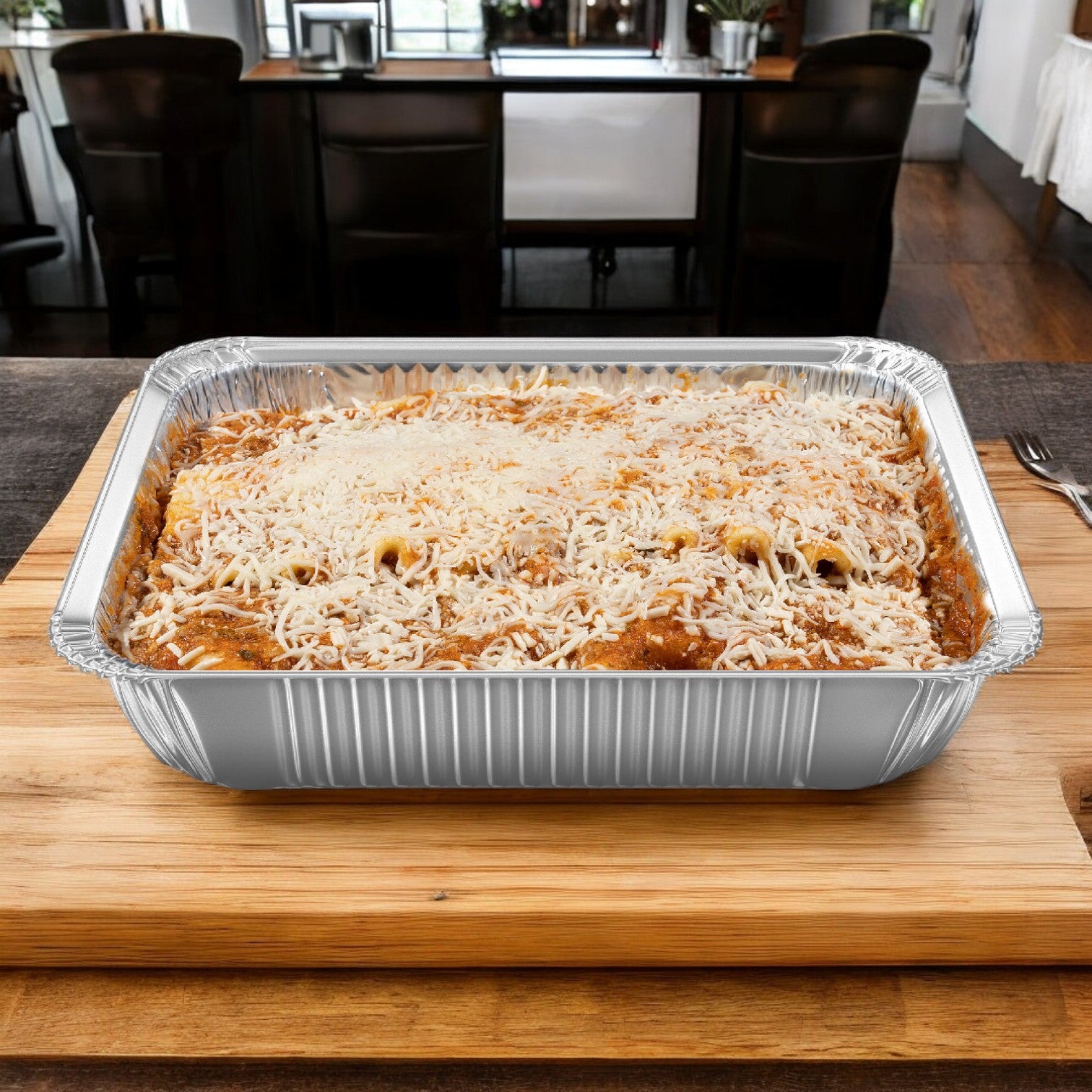 In a cozy kitchen setting, a Sysco Classic foil tray of meat lasagna from the 6 lbs. Lasagna Entree case is placed on a wooden board, topped with layers of melted cheese.