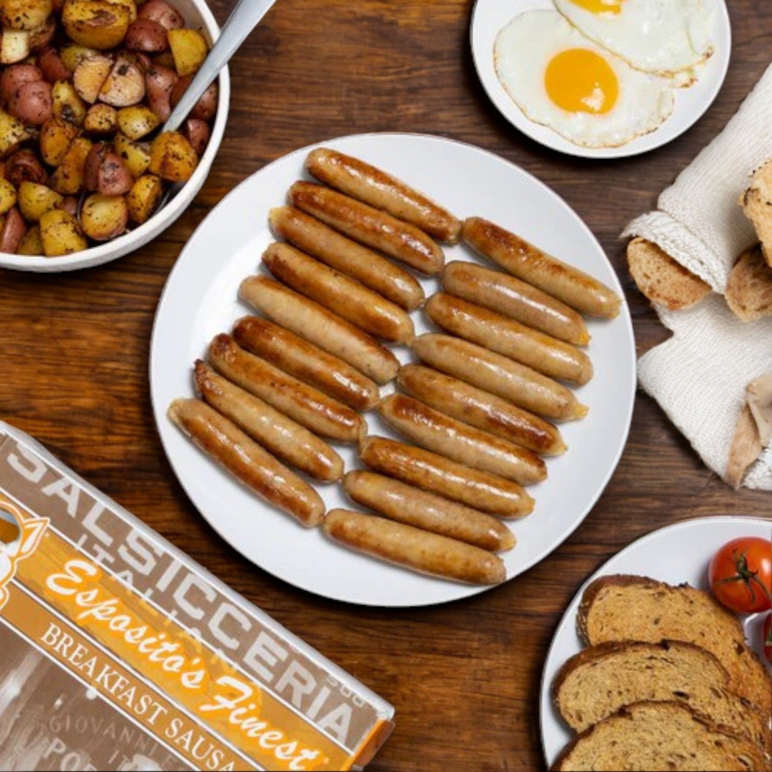 On a wooden table, a plate of cooked sausages is served with roasted potatoes, two fried eggs, sliced bread, and tomatoes. A package labeled "Esposito's Finest Quality Sausage – Breakfast Sausage" suggests premium quality pork in this authentic Italian recipe.