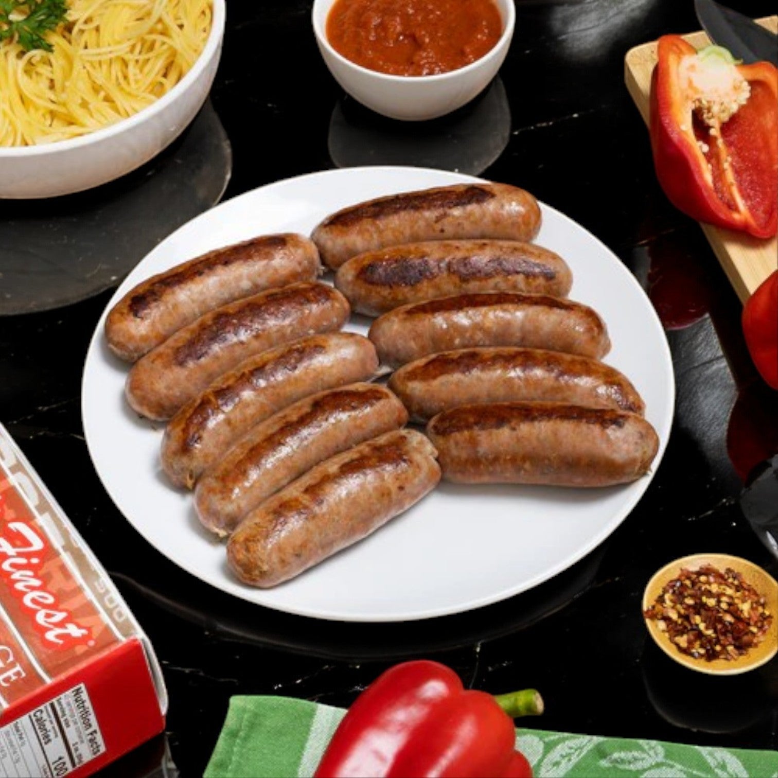 Esposito's Finest Quality Hot Italian Sausages are beautifully arranged with pasta, red bell peppers, crushed red pepper, and a bowl of sauce on a black surface, delivering an authentic Italian flavor experience.