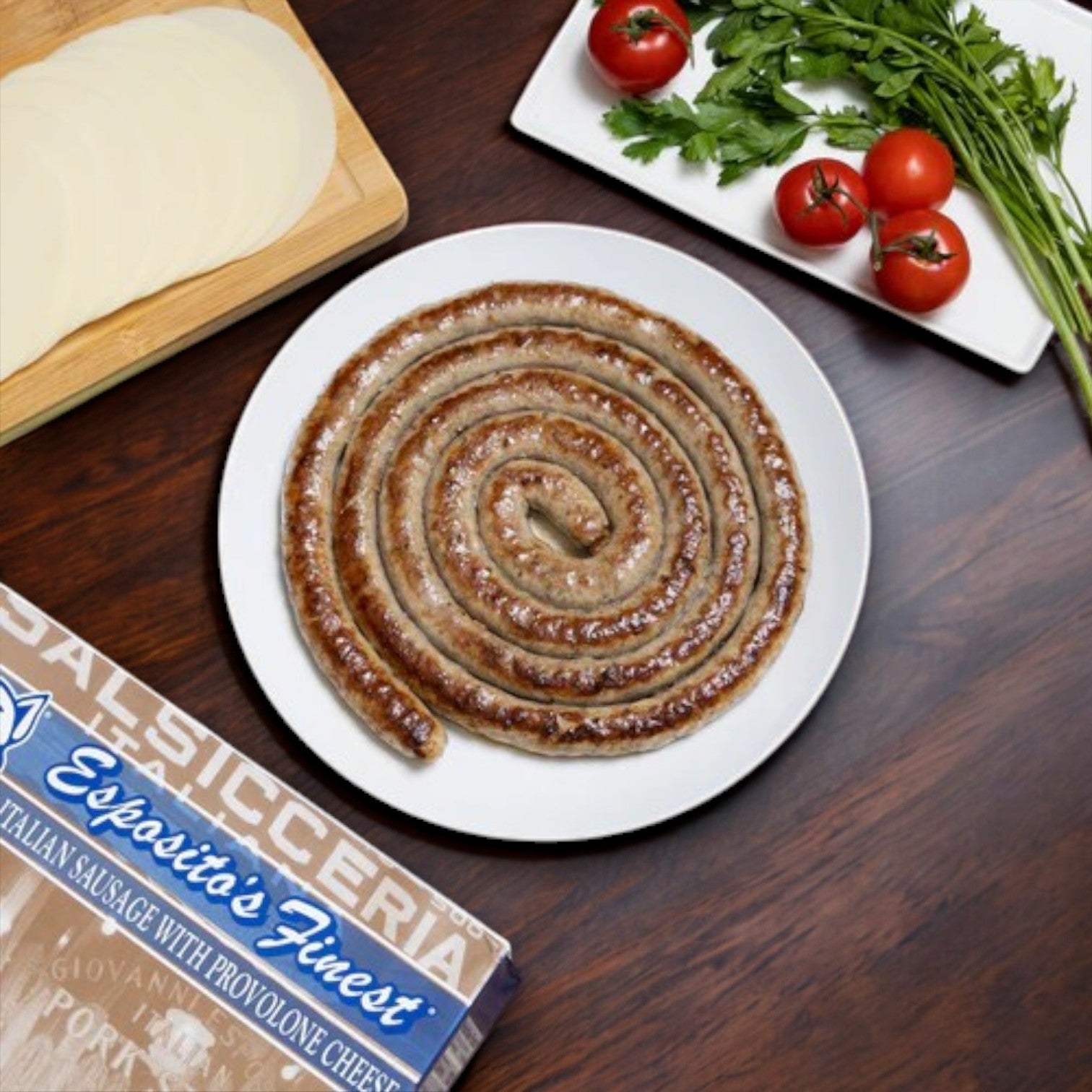 A pinwheel of Esposito's Finest Quality Sweet Sausage with Provolone and Parsley is artfully arranged on a white plate, accented by provolone slices, its box, and fresh parsley and tomatoes on a wooden table.