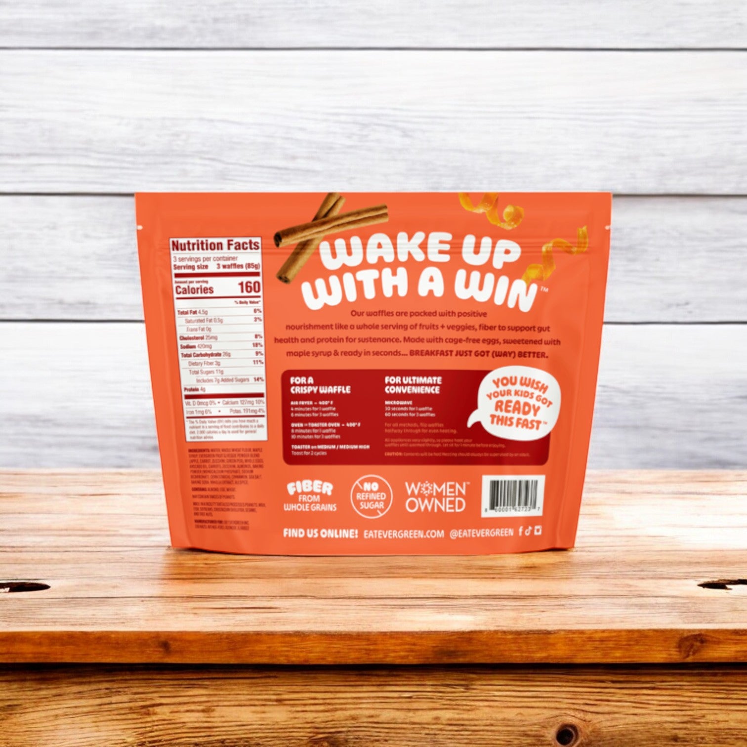 On a wooden surface lies the back view of a packaged food product. The orange package for Evergreen's Zucchini Carrot Cake Frozen Mini Waffles, 9 oz., features white and yellow text that highlights its nutritious breakfast quality, along with comprehensive nutritional information and product details.